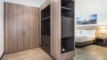 A hotel room with a large wardrobe, a bed with a headboard, a bedside table with a lamp, and a wall-mounted painting.