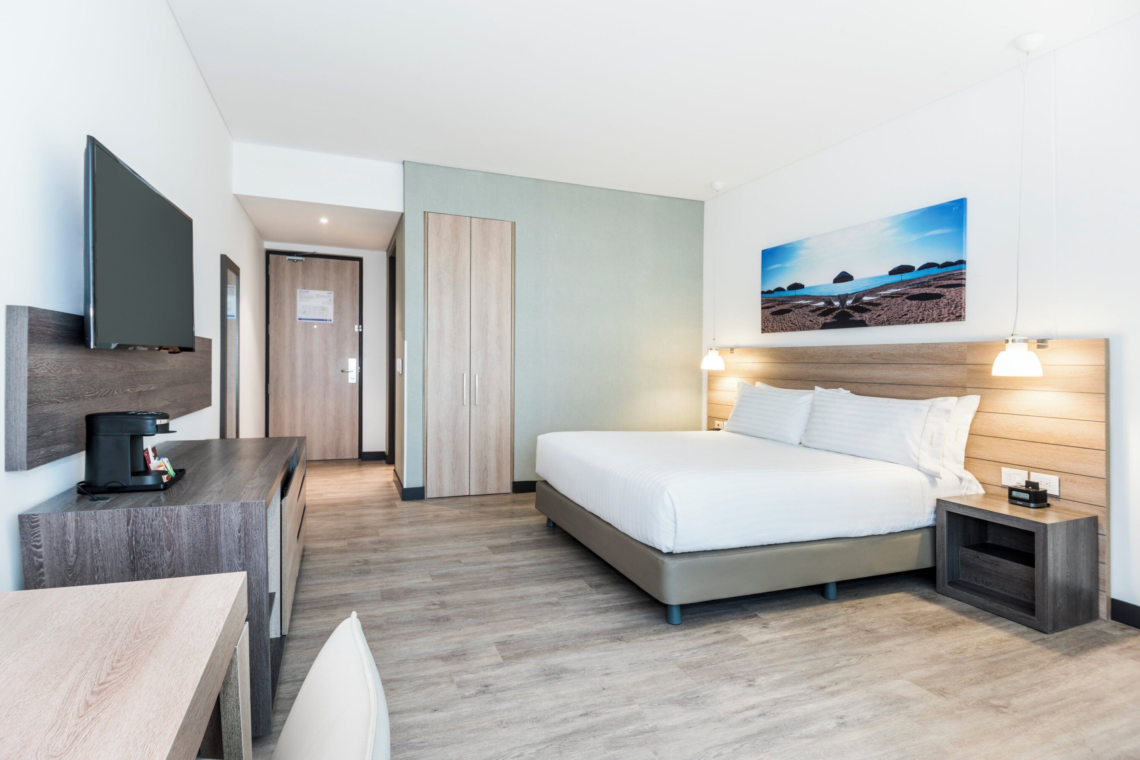 Habitación de hotel con cama, mesita de noche, televisión y un cuadro de una playa.