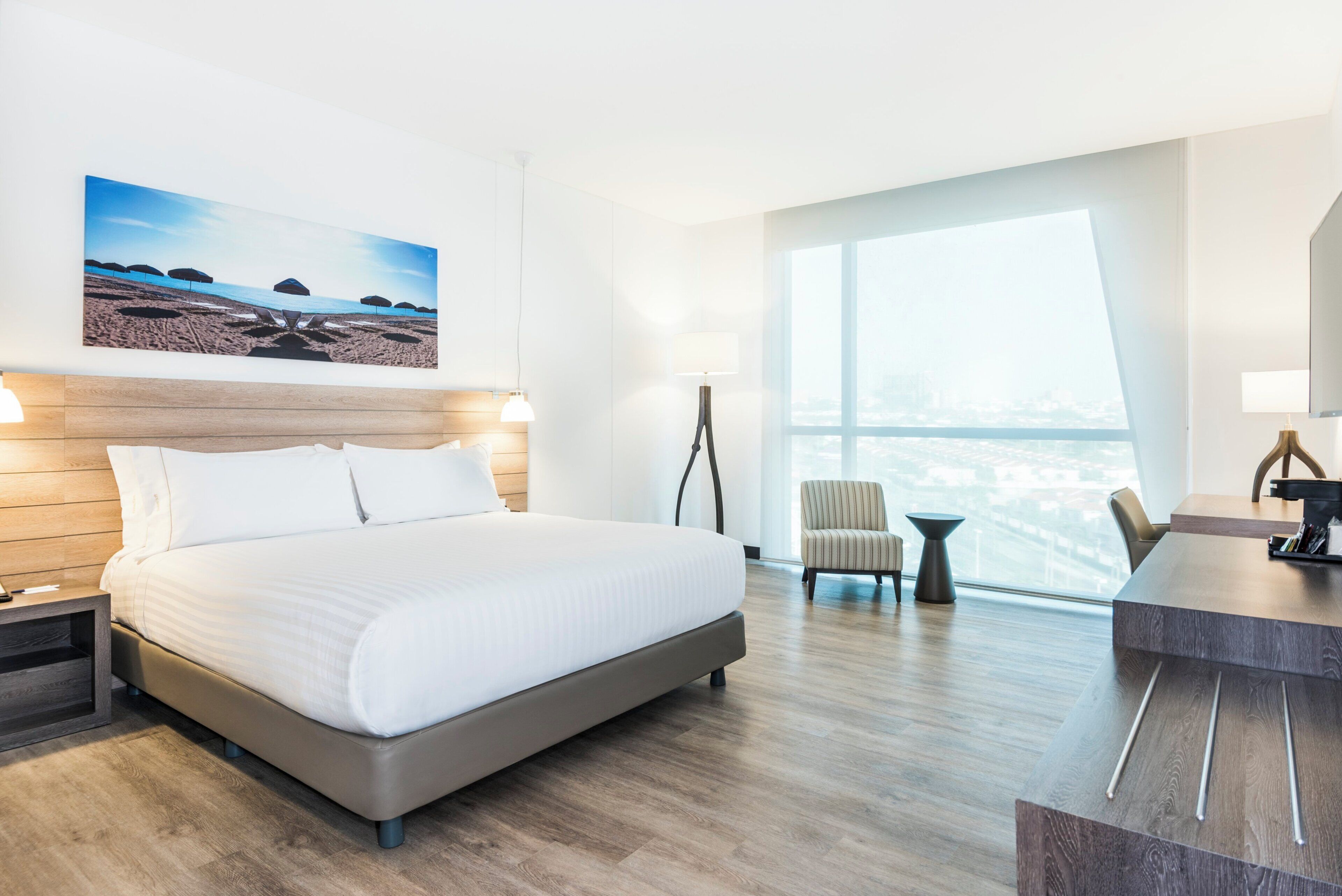Una habitación de hotel con cama, escritorio, silla, lámpara y un ventanal grande.