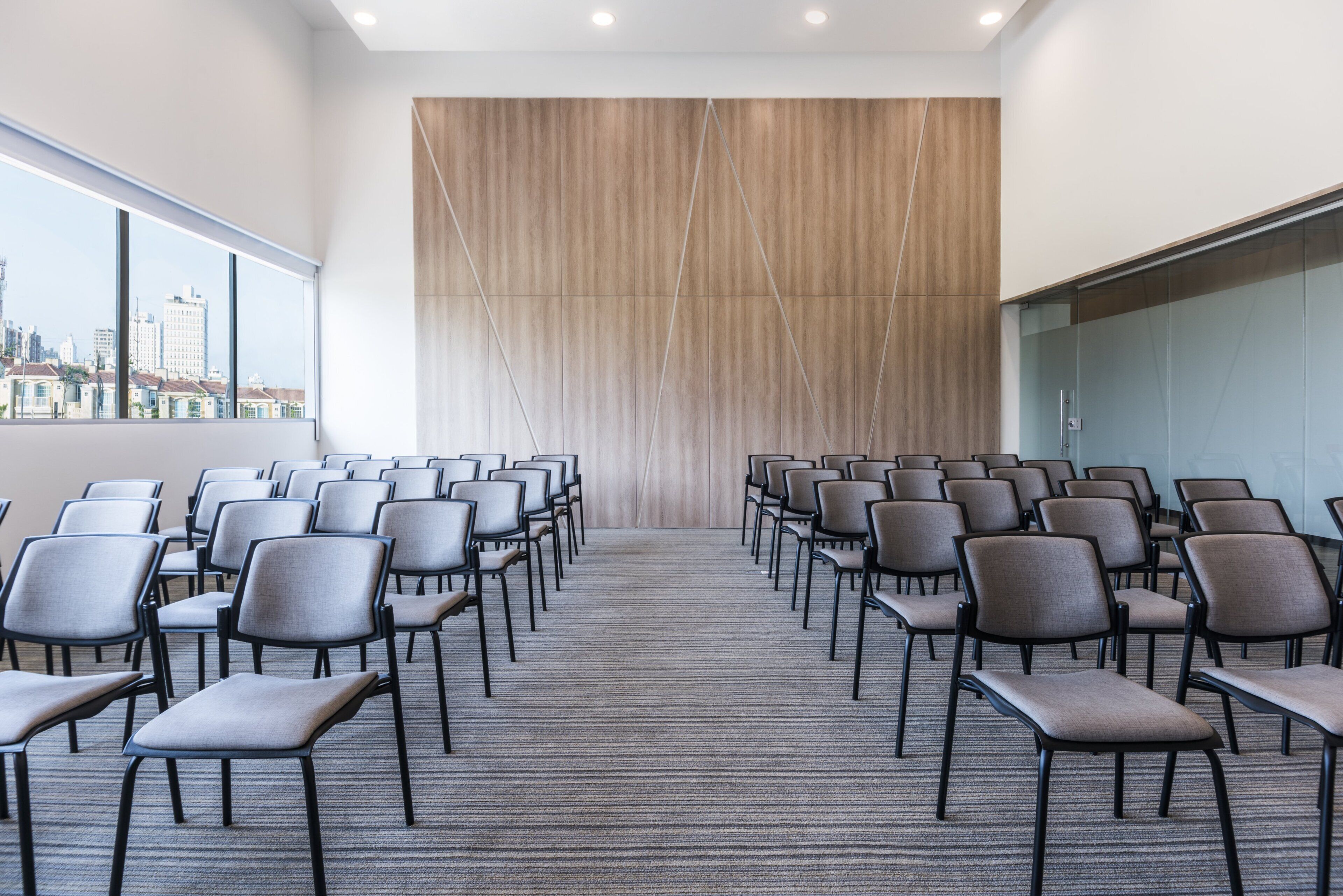 A conference room with rows of chairs facing a wooden panel wall, carpeted floor, and large windows with a city view.