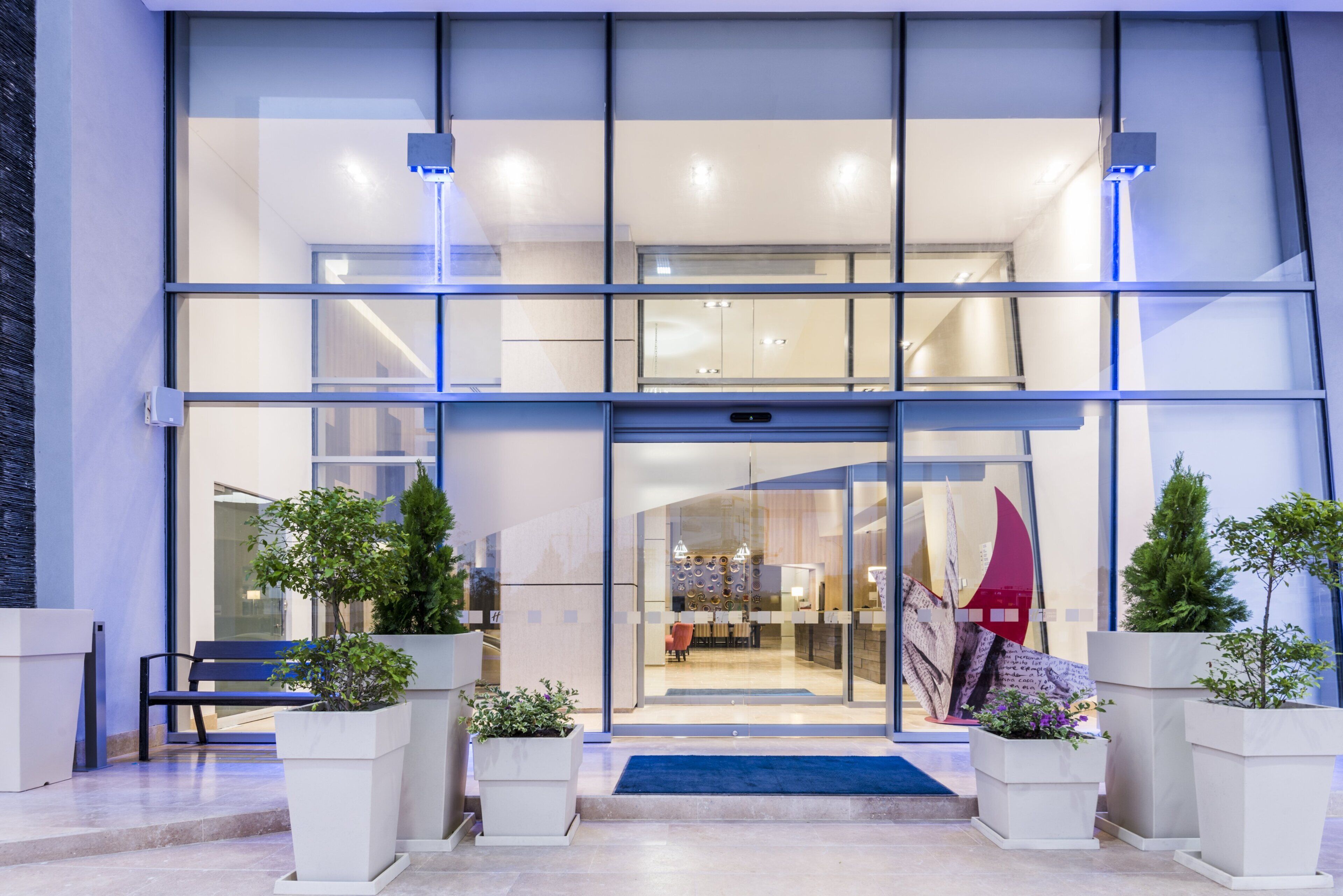 A modern building entrance with a glass facade, a reception desk, and potted plants.