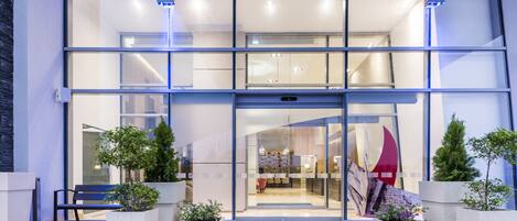 A modern building entrance with a glass facade, a reception desk, and potted plants.