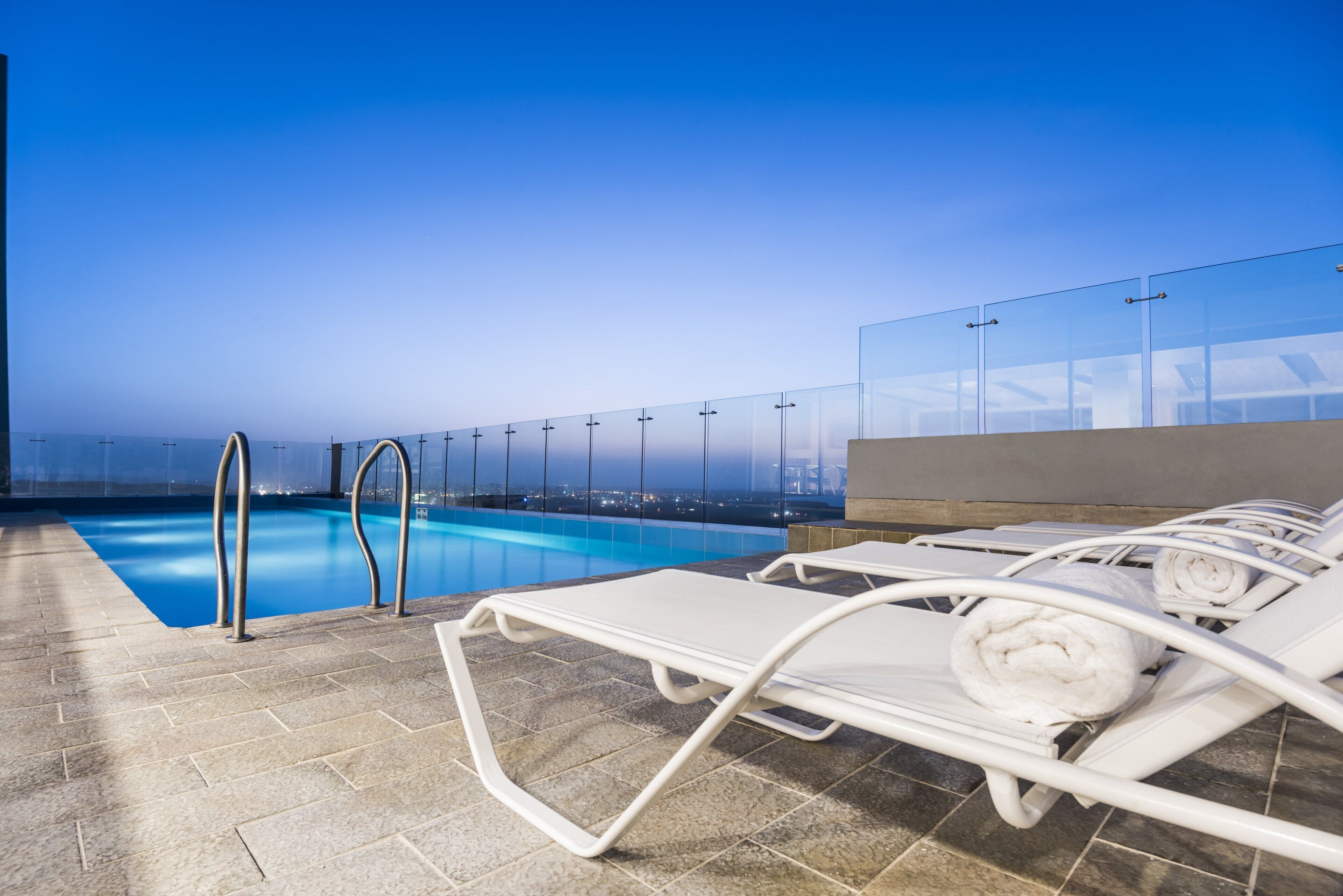 Un área de piscina en la azotea con barandillas de vidrio transparente, sillas de descanso y vistas a la ciudad al atardecer.