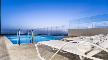 Un área de piscina en la azotea con barandillas de vidrio transparente, sillas de descanso y vistas a la ciudad al atardecer.