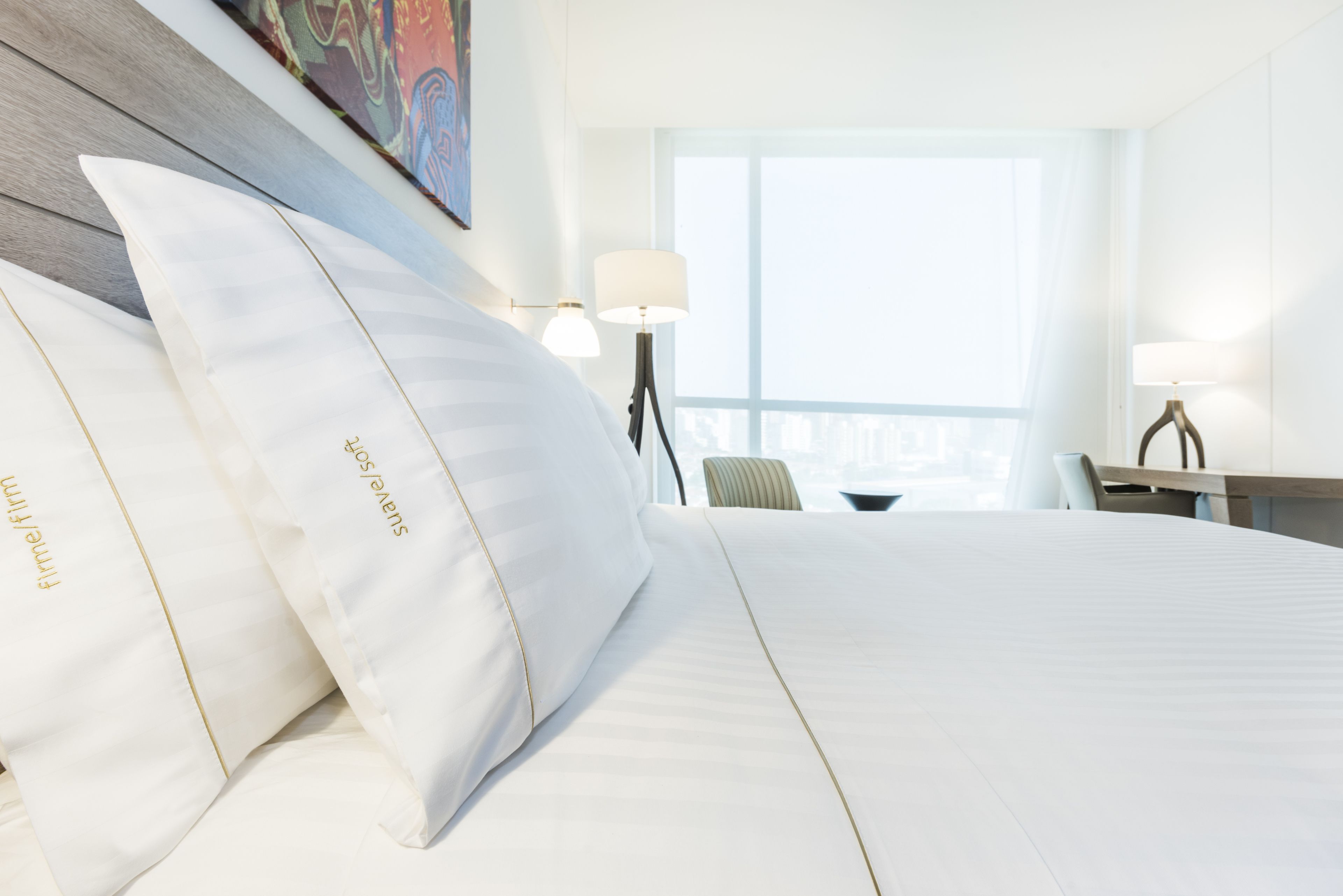 A hotel room with a large bed, two pillows, a desk, a chair, a lamp, and a window with a view.