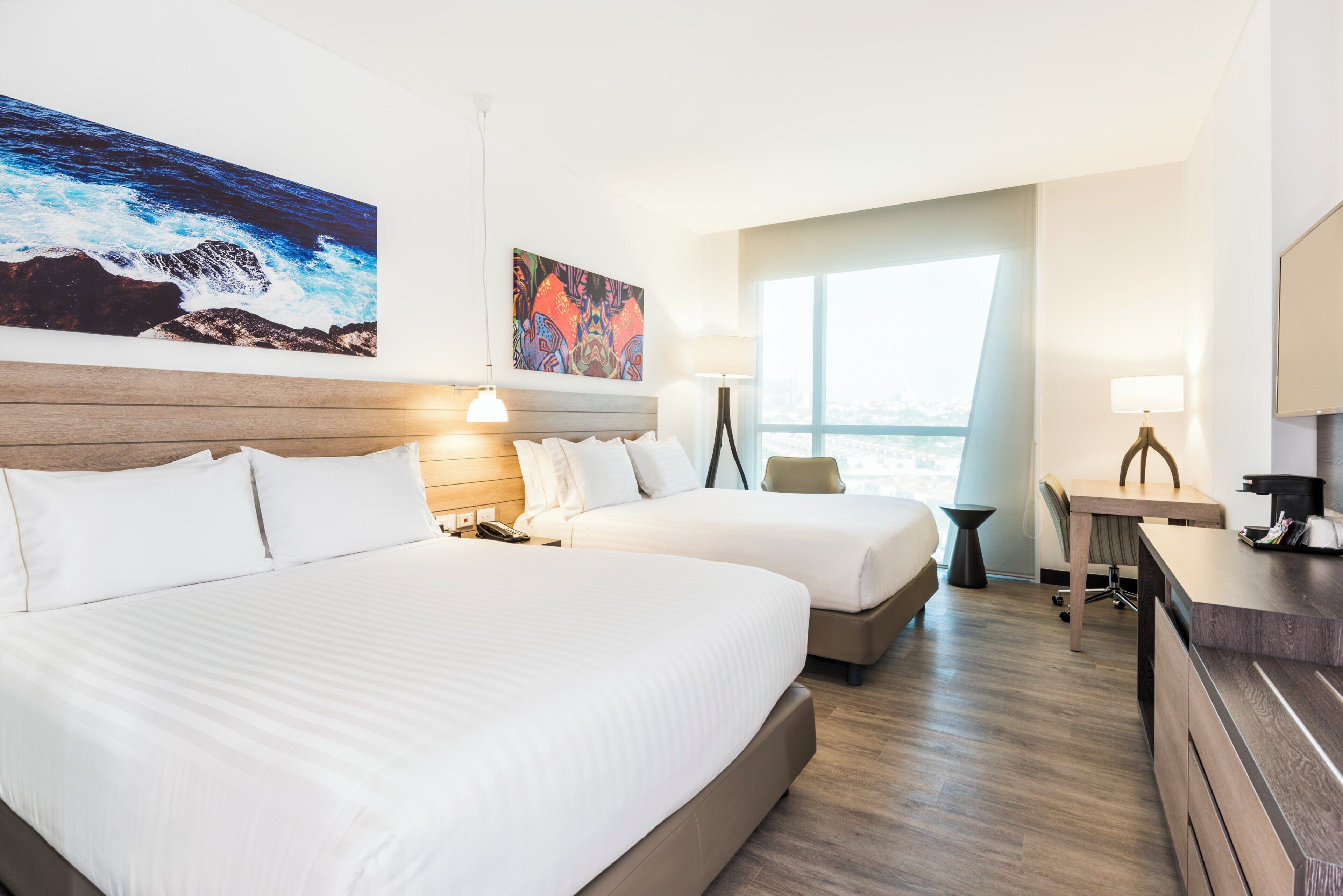 A hotel room with two beds, a desk, a chair, a large window, and artwork on the walls.