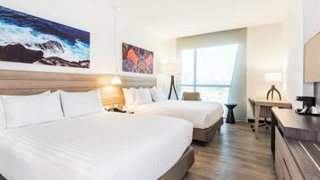 A hotel room with two beds, a desk, a chair, a large window, and artwork on the walls.