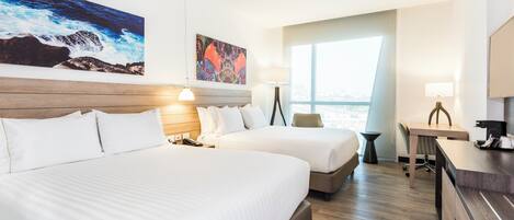 A hotel room with two beds, a desk, a chair, a large window, and artwork on the walls.