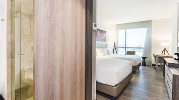 A hotel room with a glass shower enclosure, a large bed with two pillows, a desk with a chair, and a view of the city through a large window.