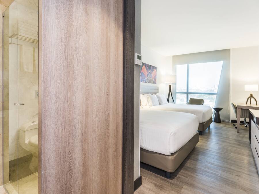 A hotel room with a glass shower enclosure, a large bed with two pillows, a desk with a chair, and a view of the city through a large window.