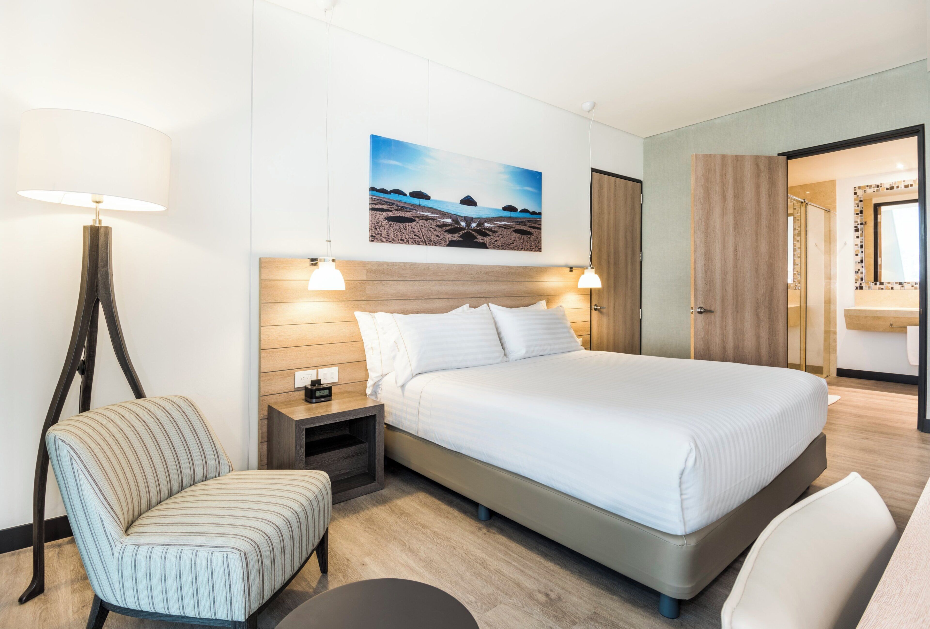 Una camera d'albergo con un letto, una sedia, un comodino con una lampada, un quadro appeso al muro e l'ingresso del bagno.