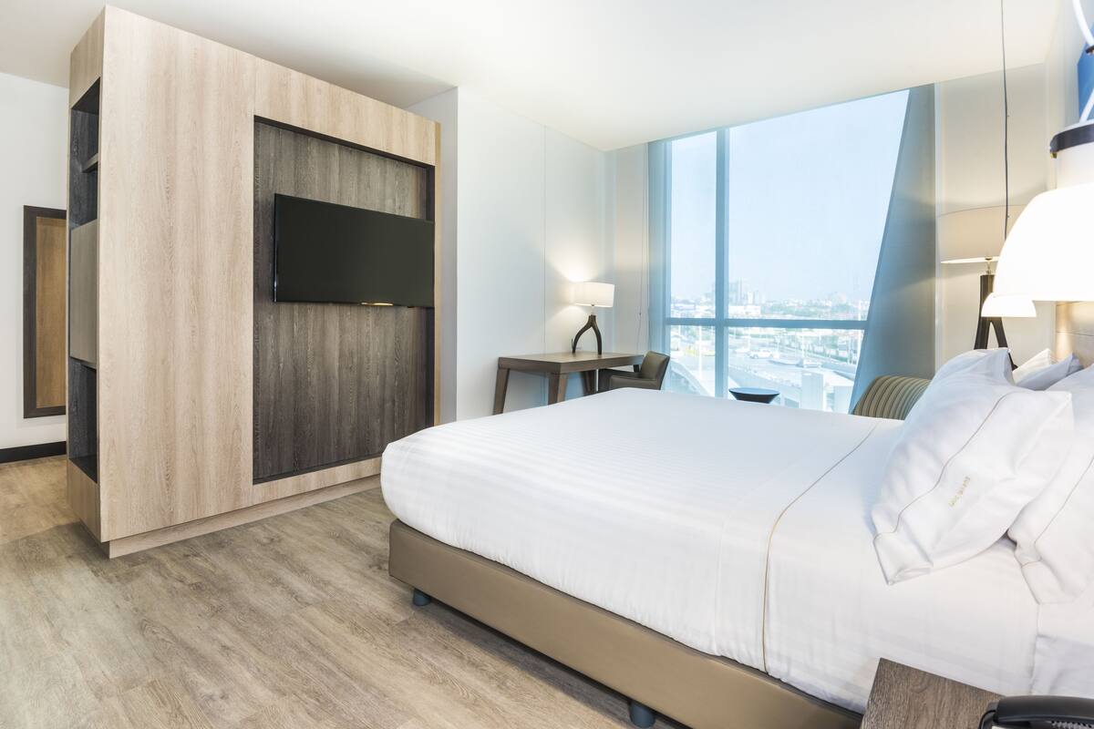 Ein modernes Hotelzimmer mit einem großen Bett, einem Flachbildfernseher an einer Holzvertäfelung, einem kleinen Schreibtisch mit Lampe und Blick auf die Stadt durch große Fenster.