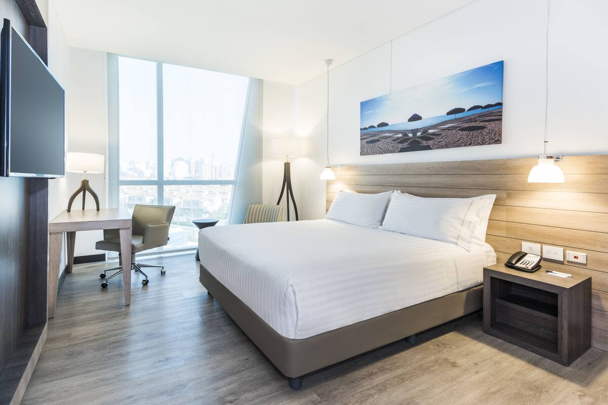 Ein Hotelzimmer mit einem großen Bett, einem Schreibtisch, einem Stuhl, einer Lampe, einem Nachttisch mit Telefon und Blick auf die Stadt.