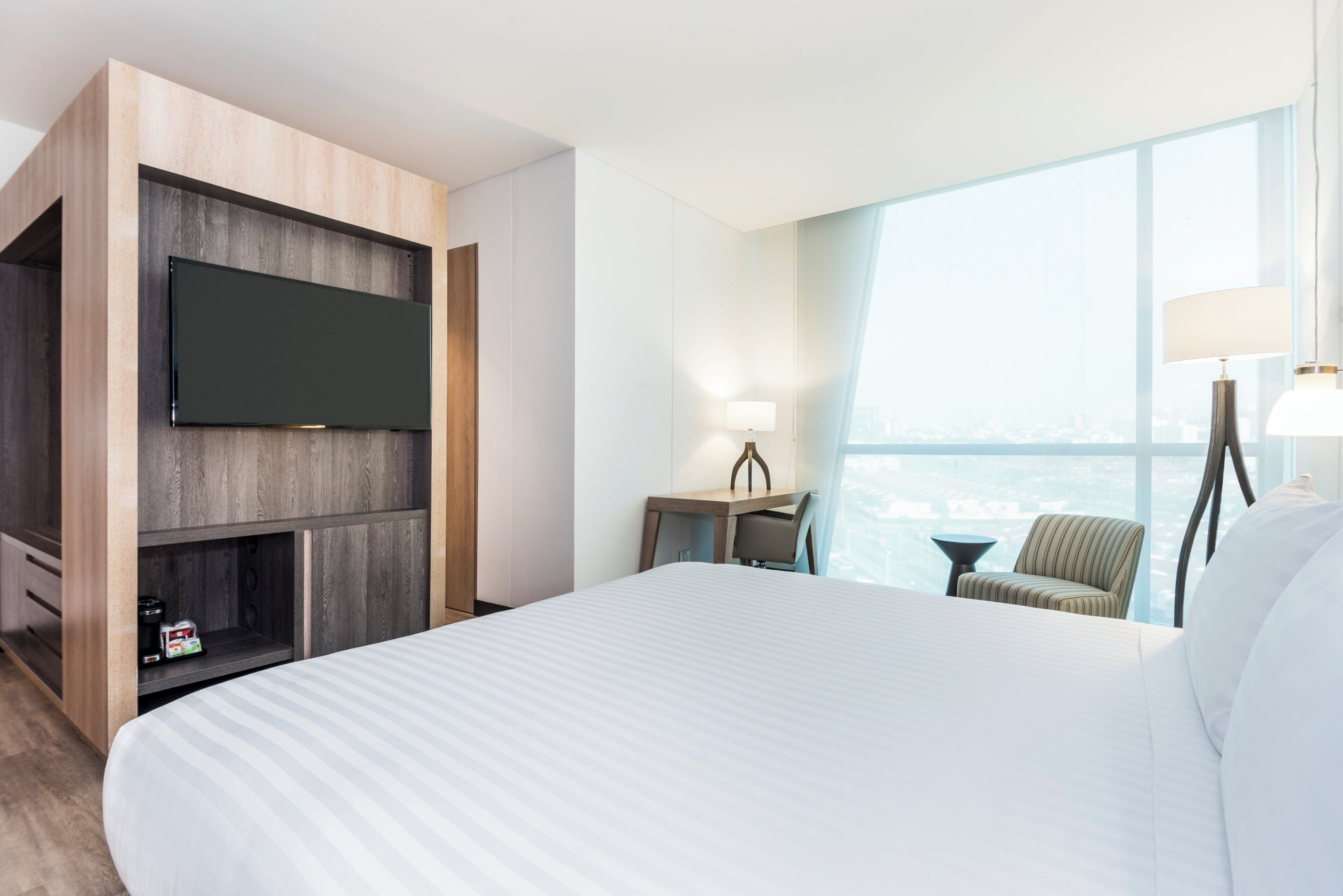 Habitación de hotel con una cama grande, un televisor de pantalla plana montado en un panel de madera, un escritorio con lámpara y vistas a la ciudad a través de amplias ventanas.