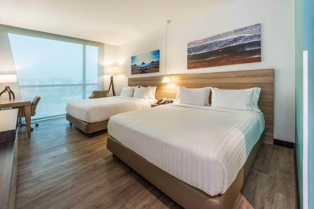 Ein Hotelzimmer mit zwei Betten, einem Schreibtisch und einem großen Fenster mit Blick auf die Stadt.