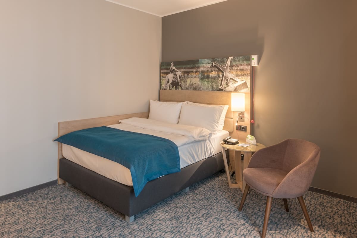 A hotel room with a bed, a chair, a nightstand, a lamp, and a painting on the wall.