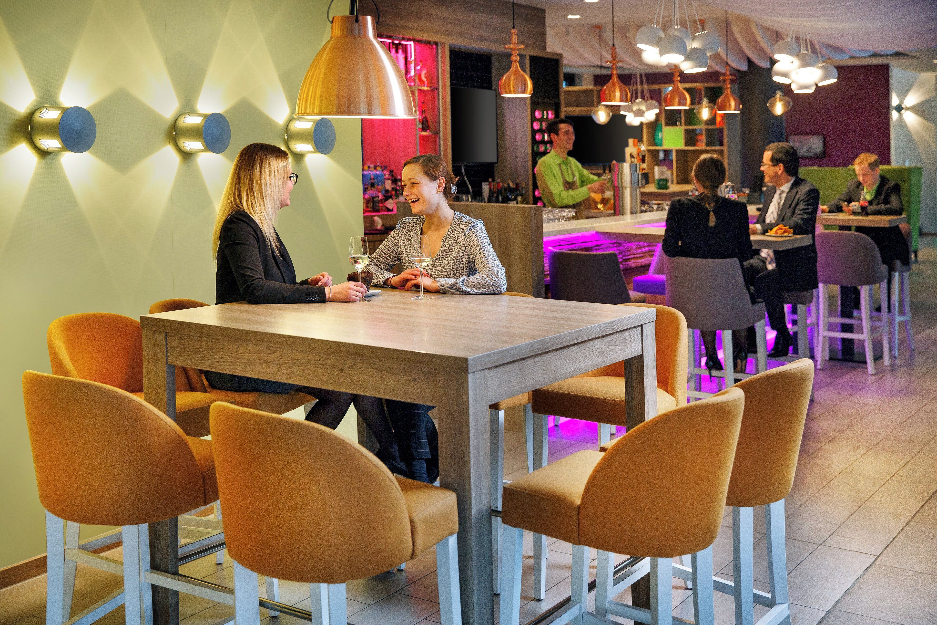 A restaurant interior with a wooden table and orange chairs, a bar area with a bartender, and hanging pendant lights.