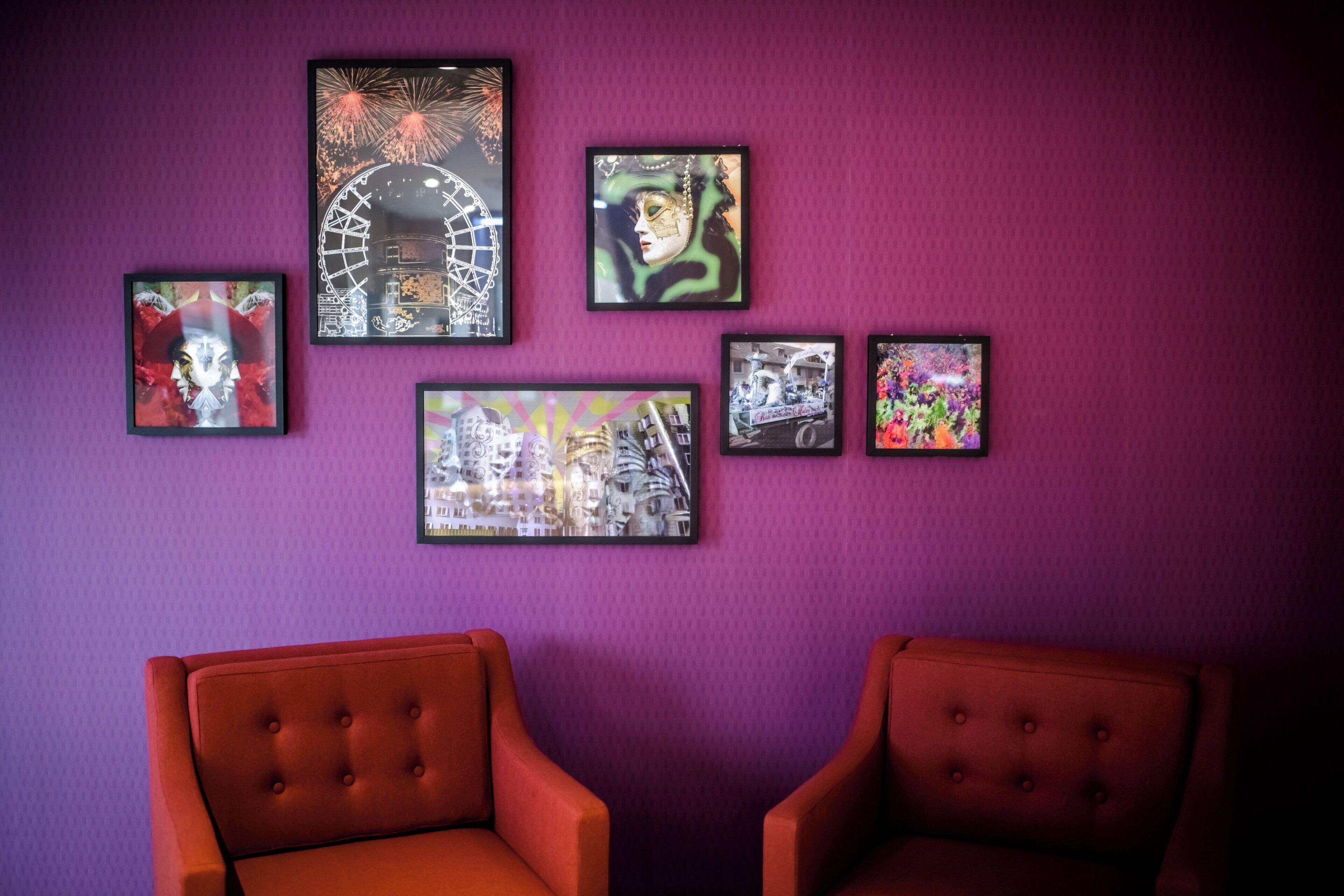 A room with a purple wall and five framed artworks. There are two red upholstered chairs.