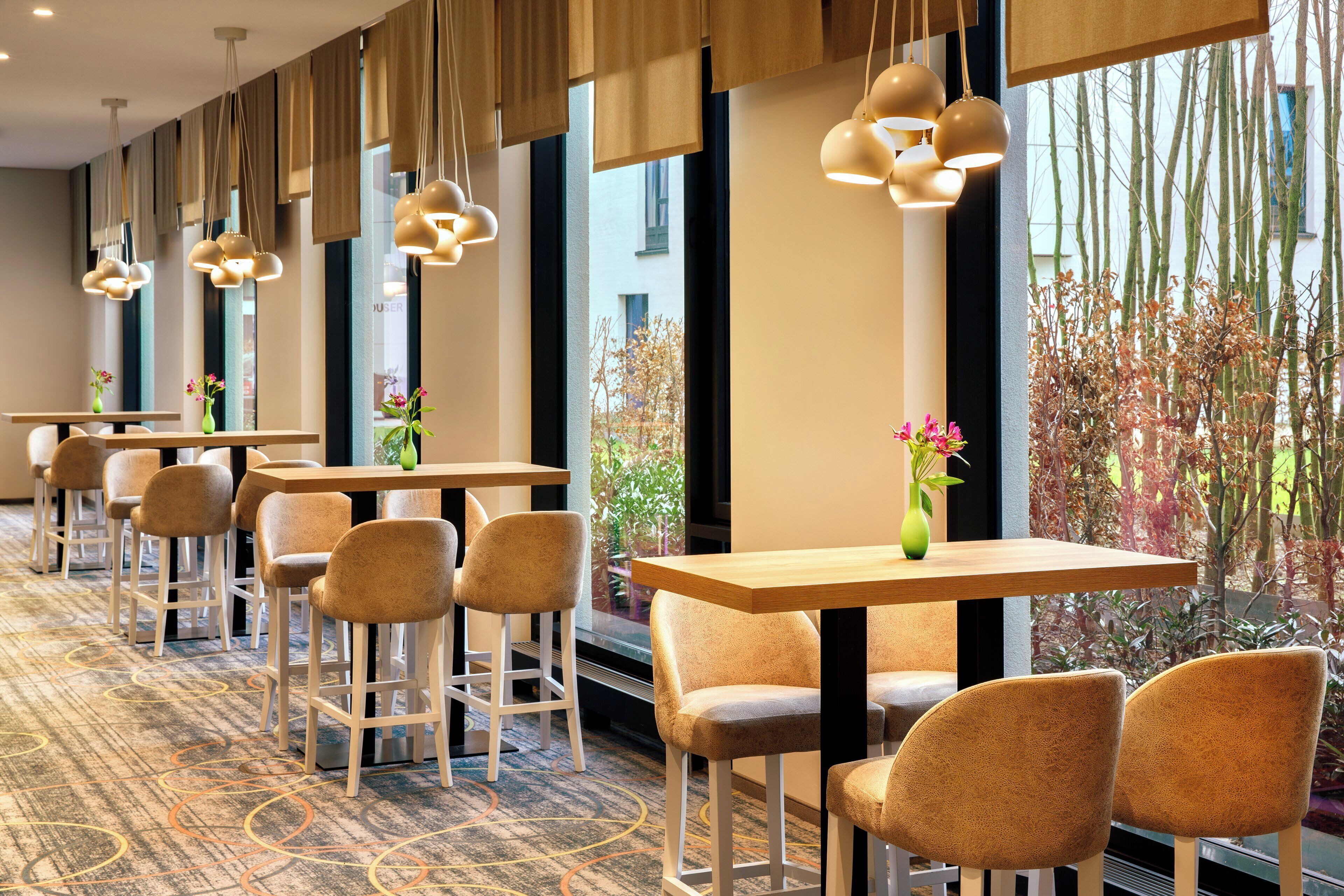 A hotel lobby with multiple tables and chairs, large windows with views of trees, and hanging pendant lights.