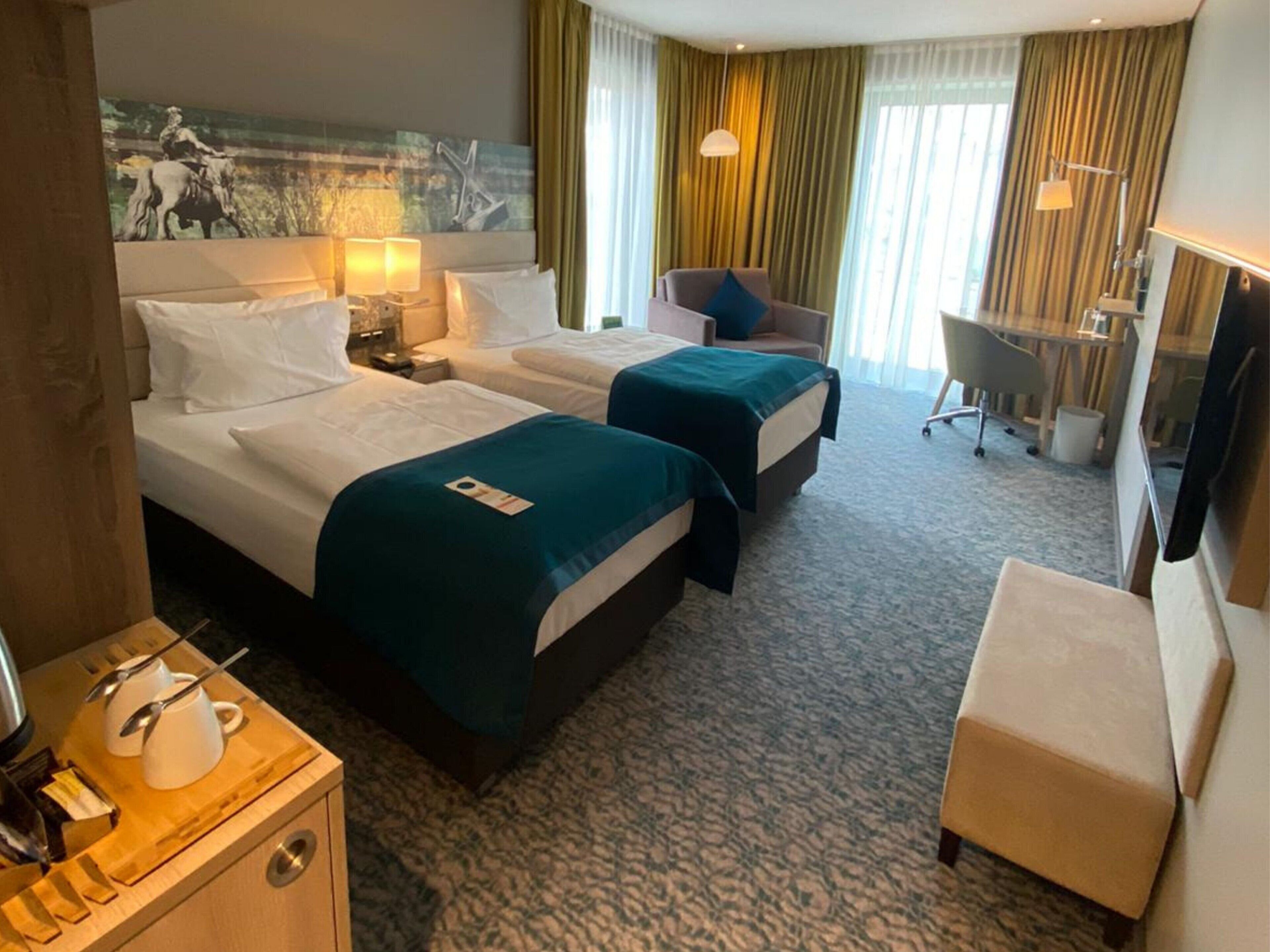 A hotel room with two beds, a desk, a chair, a TV, and a painting on the wall.