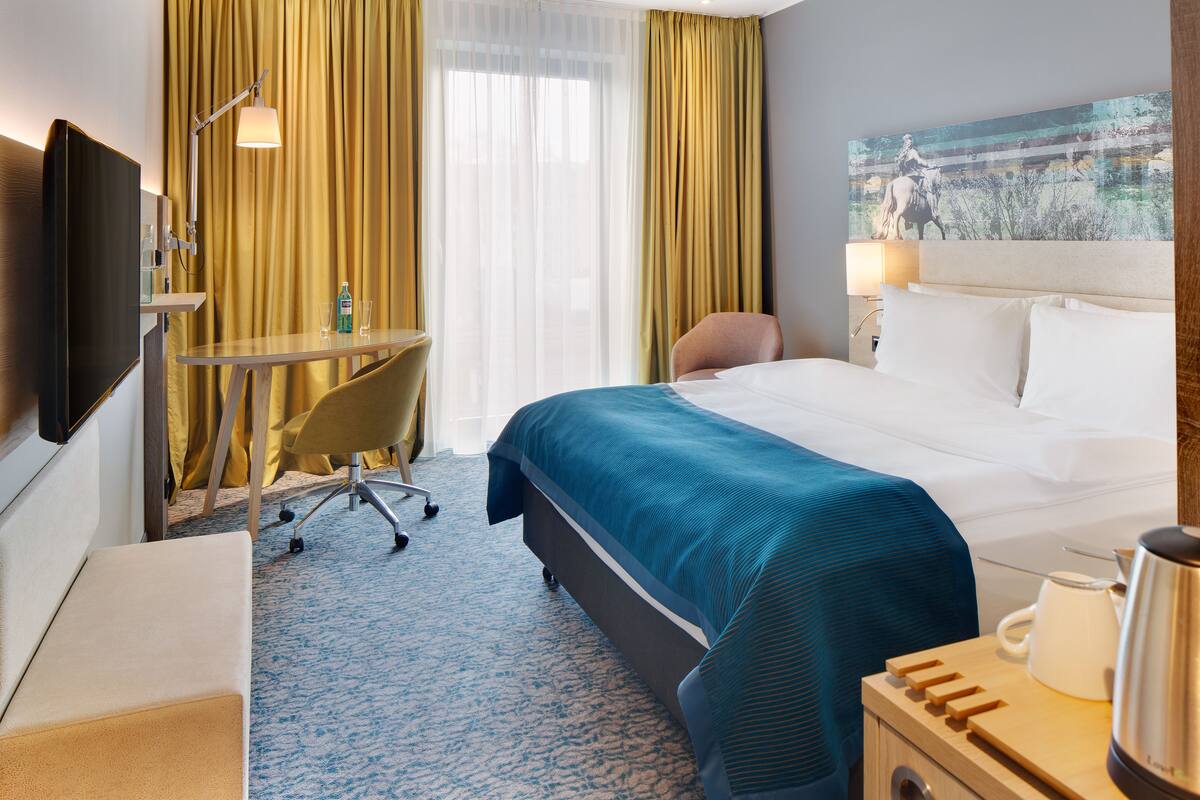 A hotel room with a bed, a desk with a chair, a television, a window with curtains, and a painting on the wall.