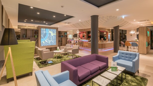 Un hall d’hôtel moderne doté d’espaces aménagés avec des sièges, d’un bar et d’un grand écran affichant une carte numérique.