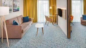 A hotel room with a sofa, a small table, a chair, a TV, and artwork on the wall.