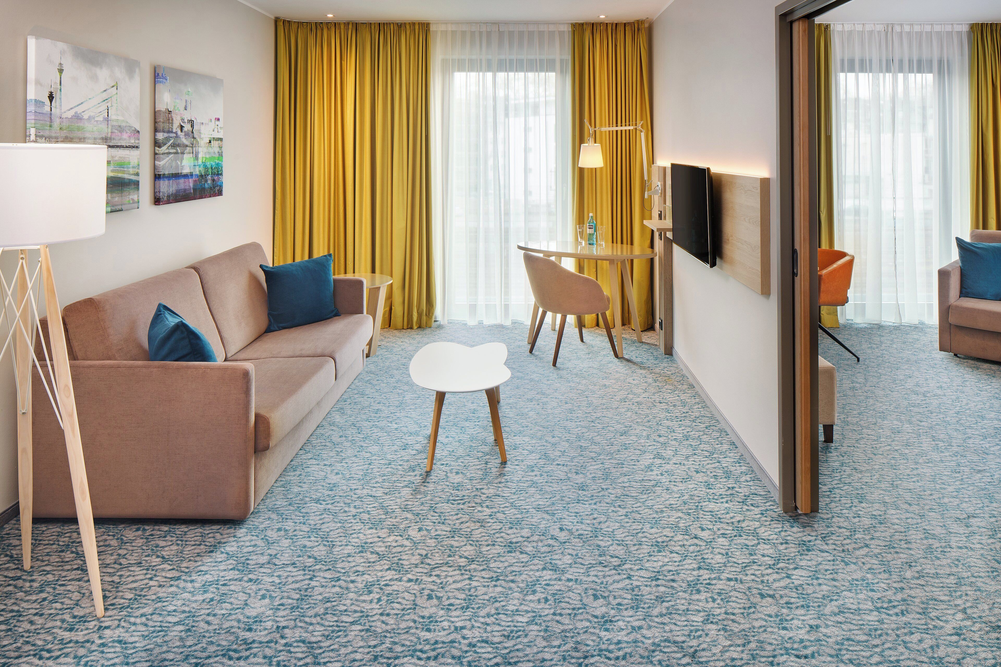 Hotellihuoneessa on sohva, pieni pöytä, tuoli, seinälle asennettu televisio ja suuri ikkuna, jossa on verhot.
