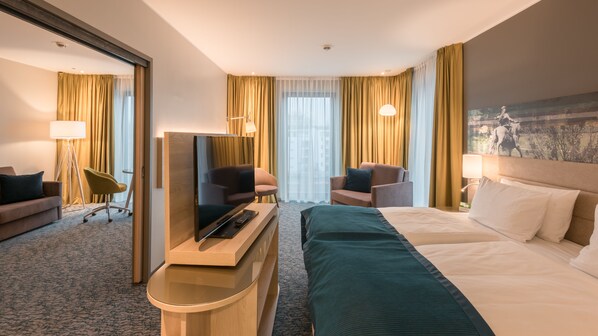 Habitación de hotel con cama, dos sillones, escritorio con televisor y ventana grande con cortinas.