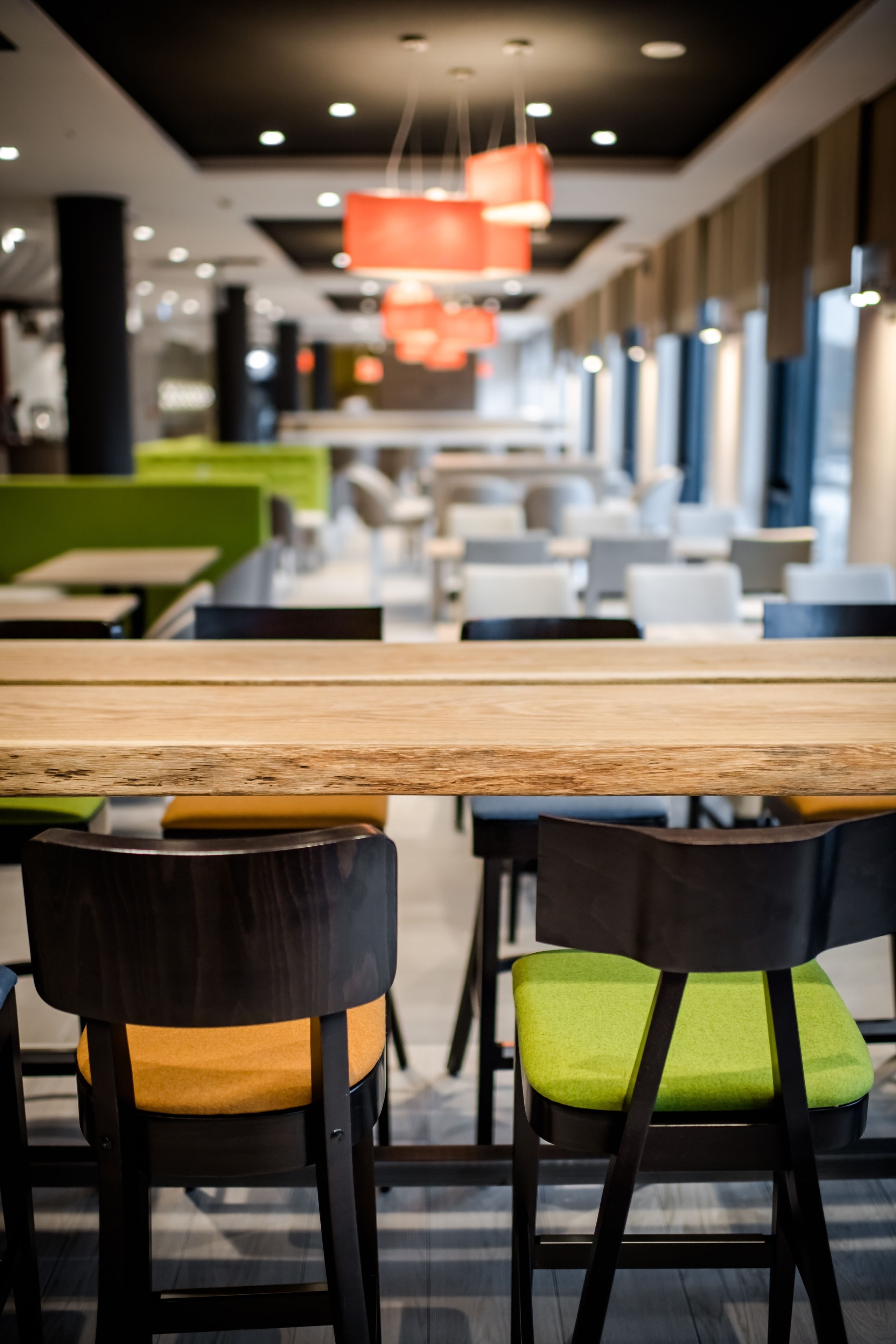 Un comedor moderno con una mesa de madera larga, sillas negras con cojines verdes y lámparas colgantes naranjas.