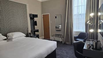 Habitación de hotel con una cama grande, un escritorio con silla, un espejo con luces, una ventana con cortinas y un radiador fijado a la pared.