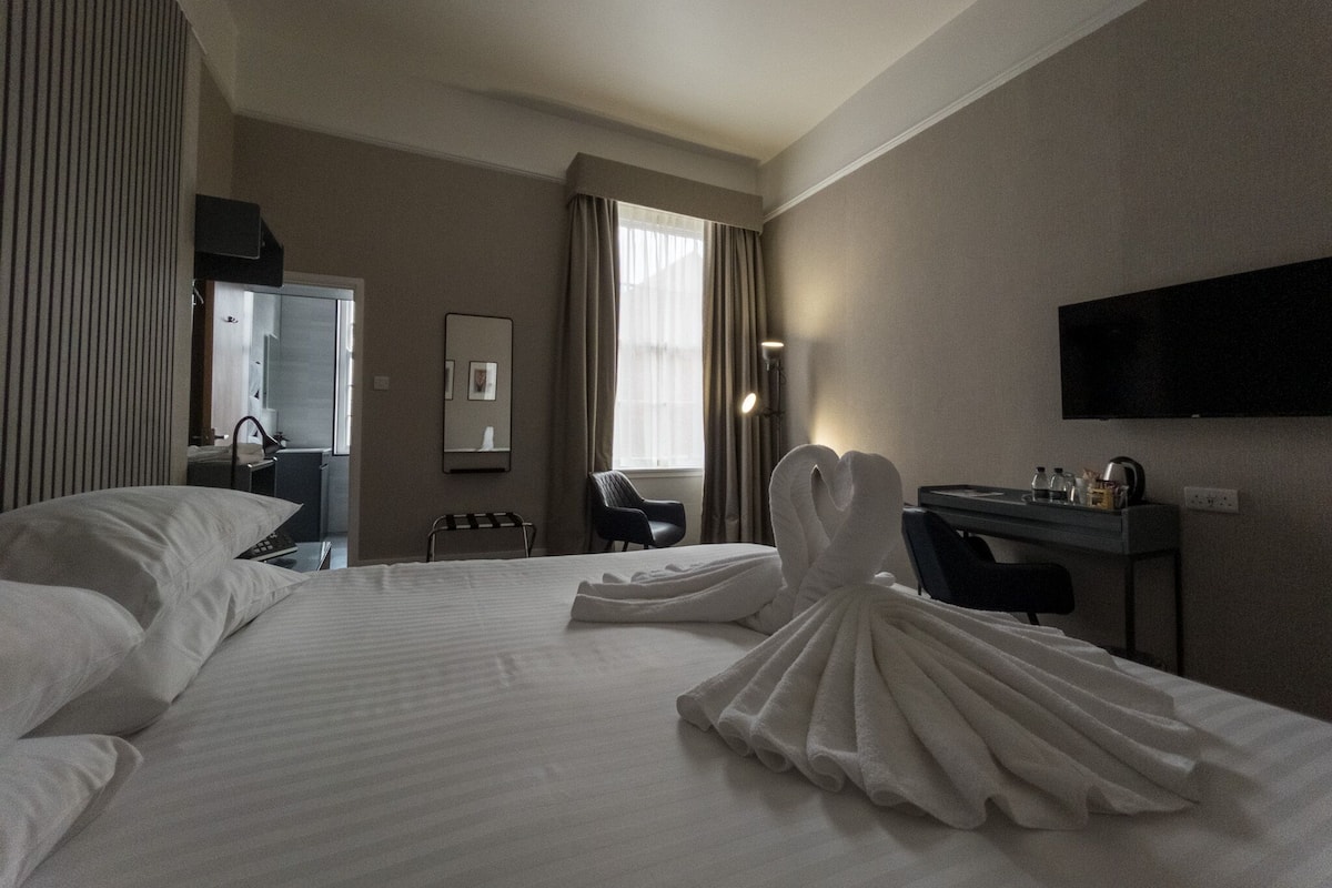 Ein Hotelzimmer mit einem großen Bett, einem Schreibtisch, einem Sessel, einem Flachbildfernseher und einem Fenster mit Vorhängen.
