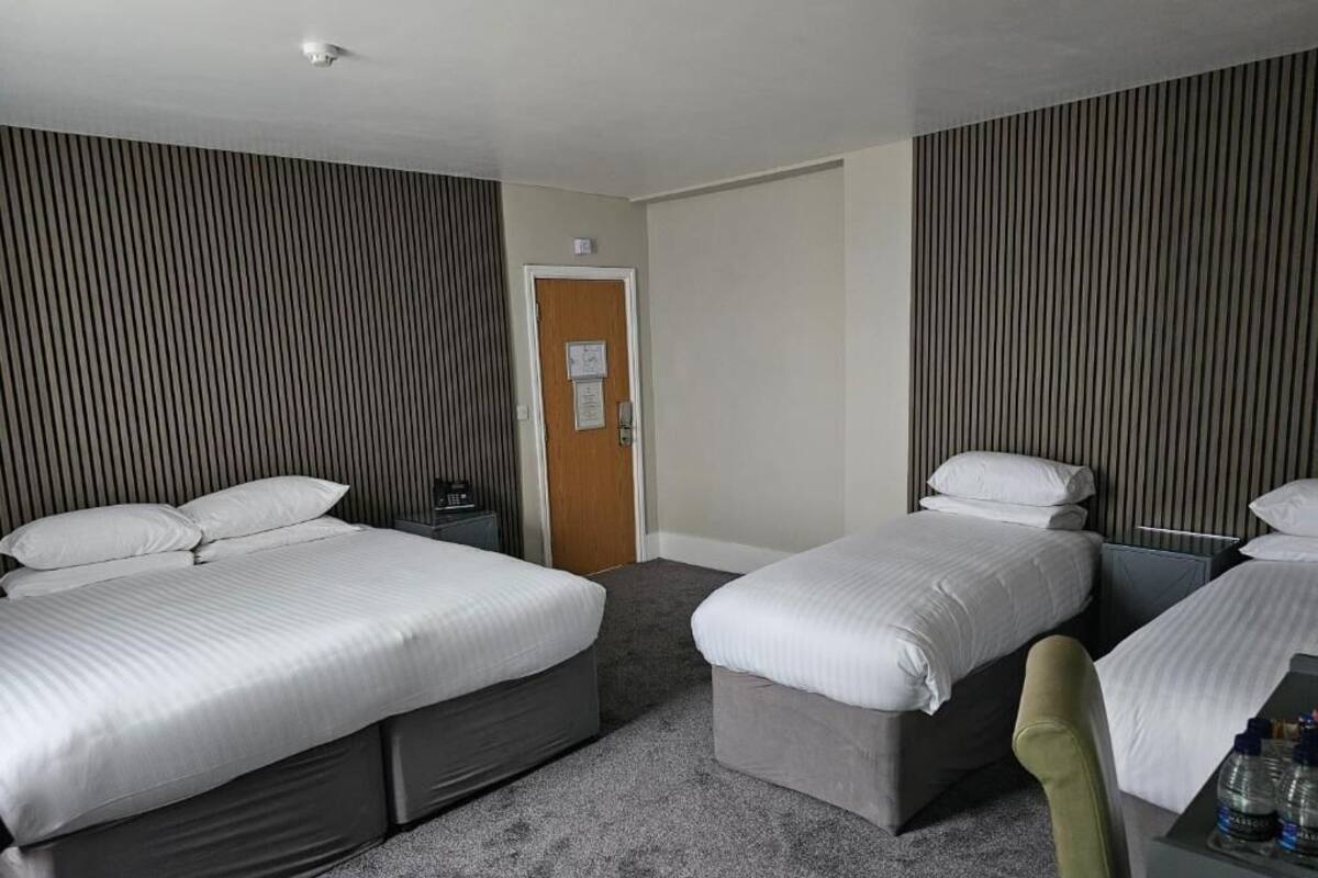 Ein Hotelzimmer mit zwei Einzelbetten, einer Holztür, einem kleinen Tisch mit Stuhl und einem grauen Teppich.