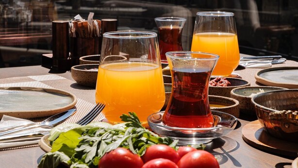 Una mesa dispuesta con vasos de jugo de naranja, un vaso de té y un plato de tomates, con una vista de la montaña al fondo.