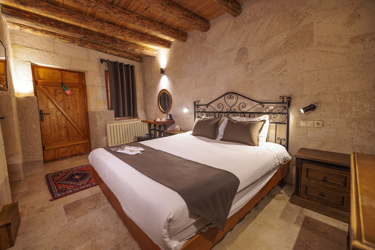Un dormitorio con una cama de madera, una mesita de noche, un aparador, un muro de piedra, una puerta de madera y una pequeña mesa con una lámpara.