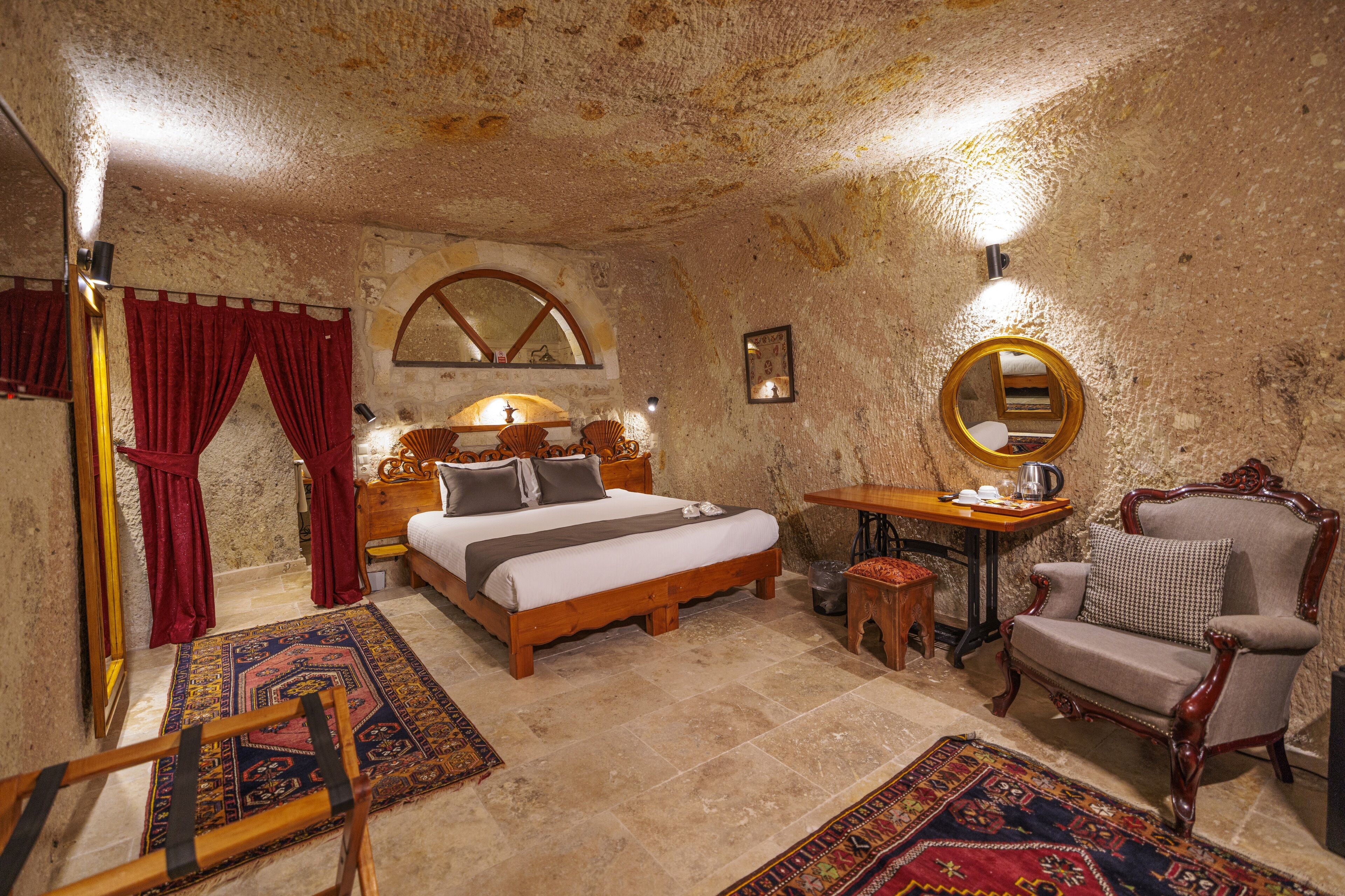 A rustic bedroom with a large bed, a wooden nightstand, a small table, a chair, and a mirror.
