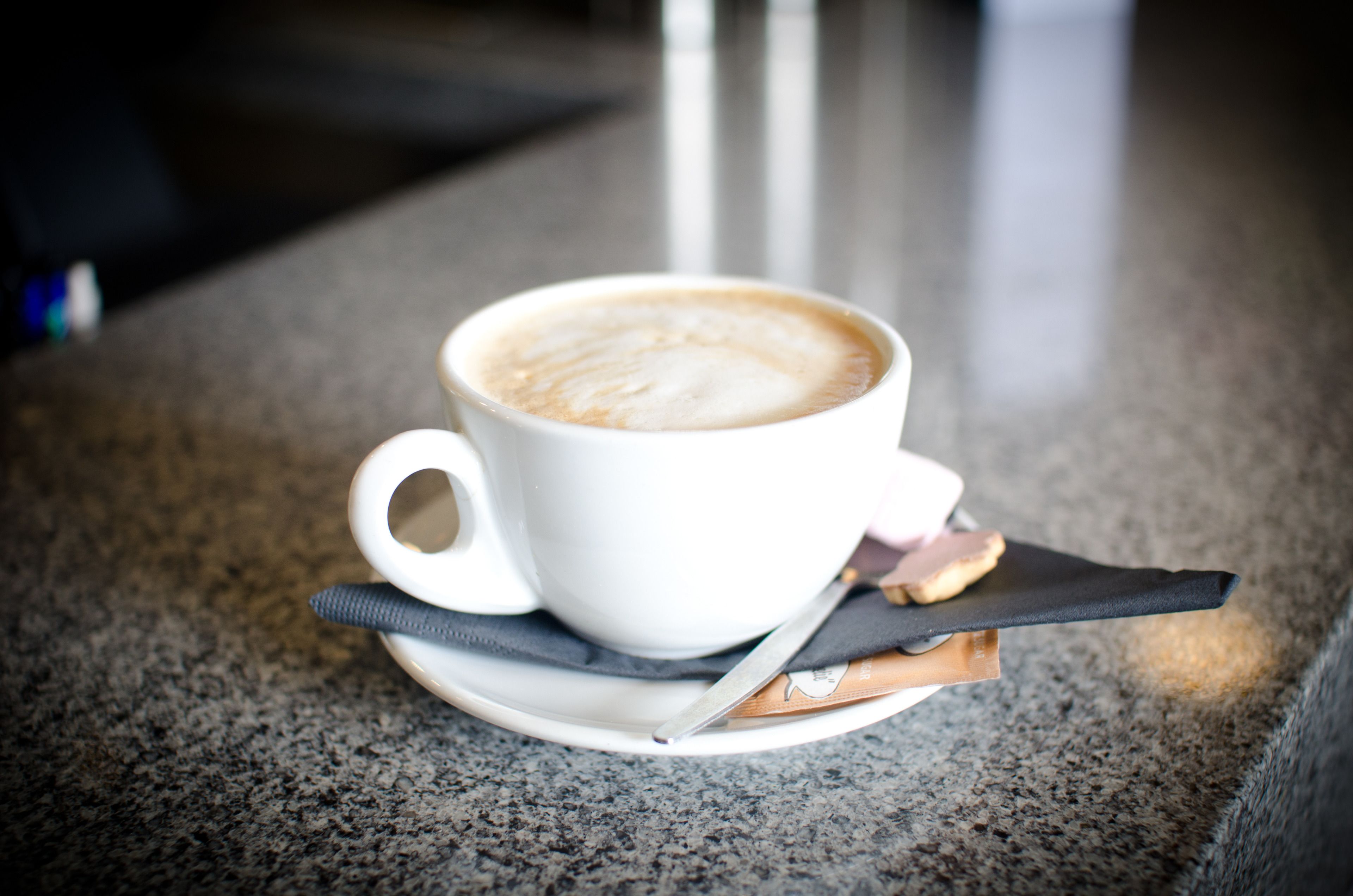 A white cup of coffee on a saucer with a spoon and a napkin on a granite countertop.
