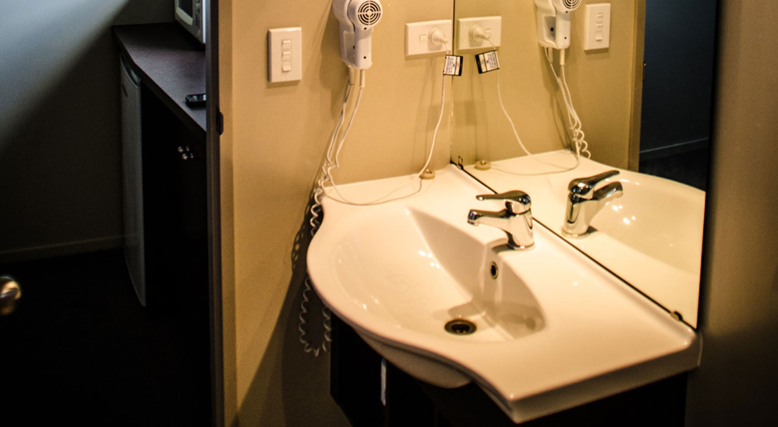 A bathroom with a white sink, two faucets, and a mirror. There are hair dryers mounted on the wall.