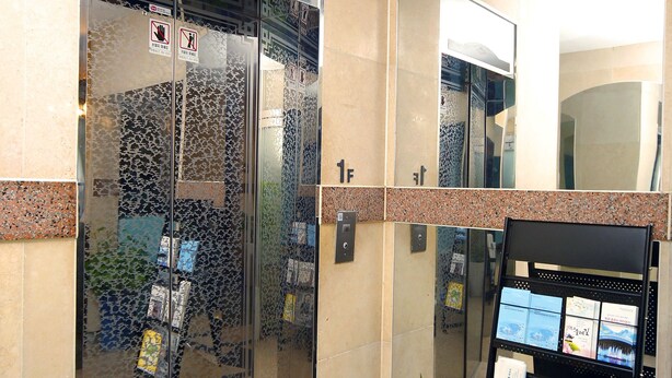 An elevator with glass doors, a small plant on a stand, and a magazine rack with brochures. - Ekonomy Hotel Mokpo (Mokpo)