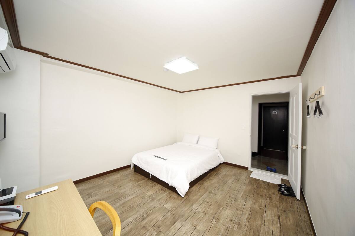 Un dormitorio con cama, escritorio y silla, una mesita y una puerta, además de una lámpara en el techo.