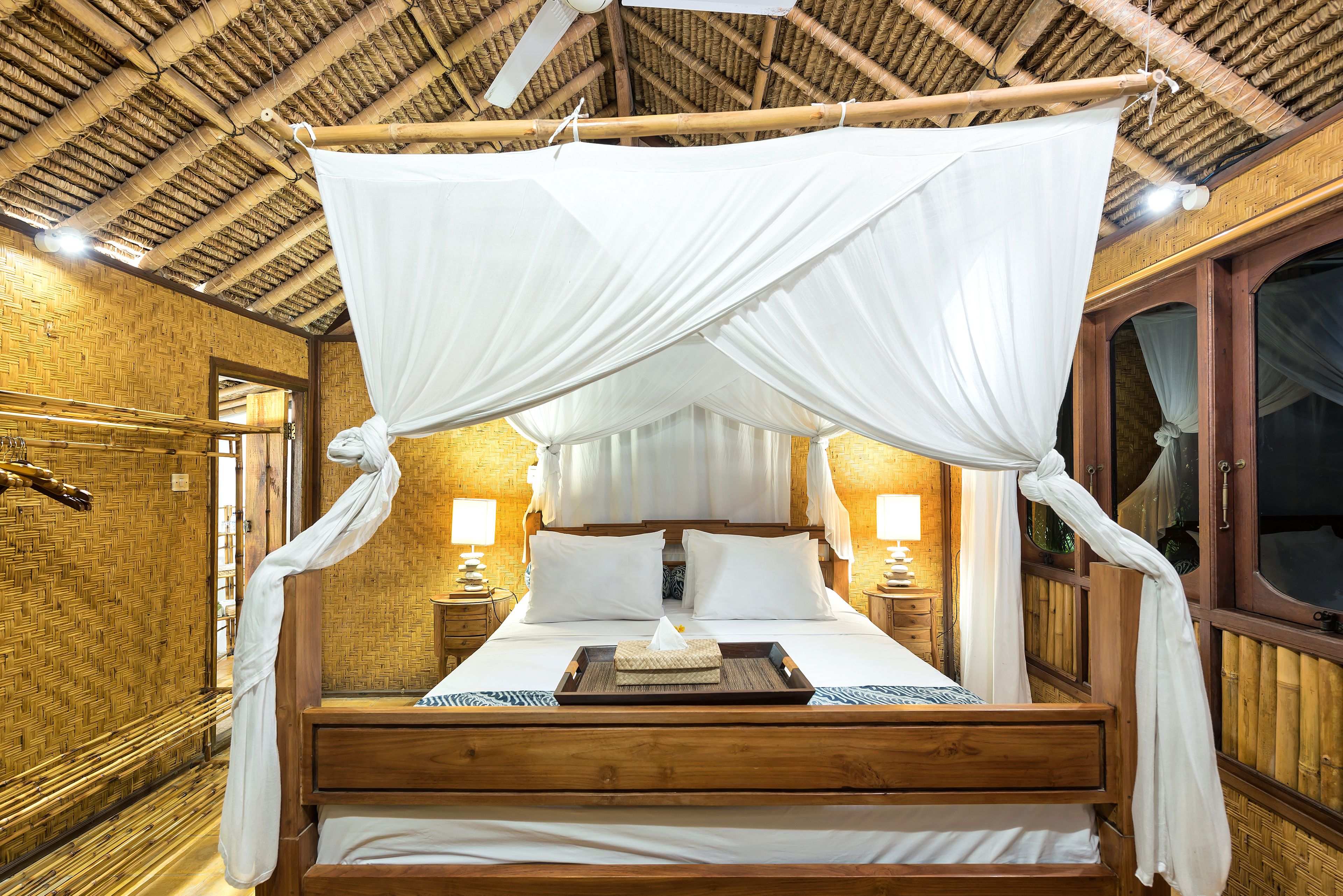 Una cama con dosel y cuatro columnas, con sábanas blancas, cabecero de madera y lámparas a cada lado.