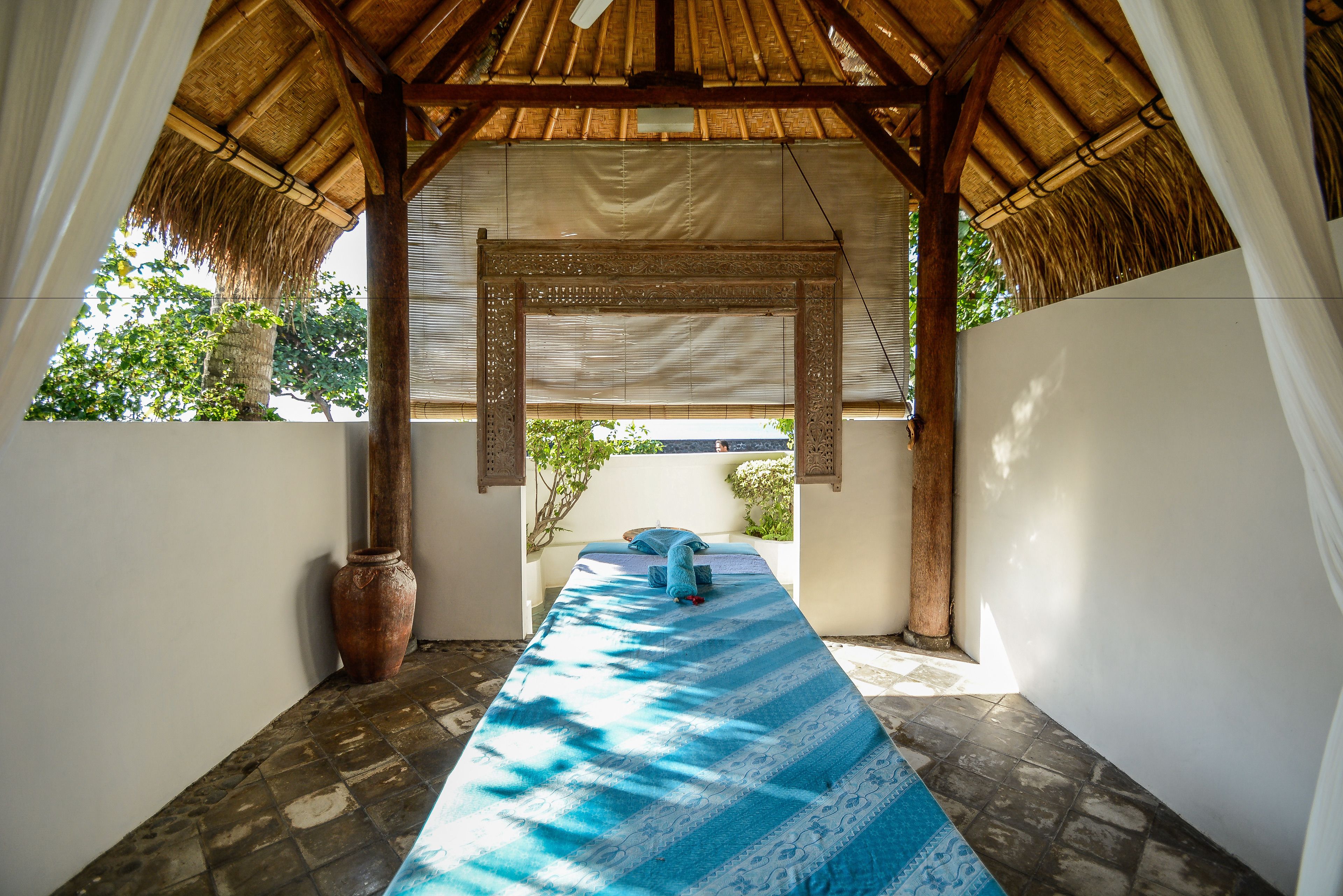 A room with a thatched roof, a bed with a blue patterned bedspread, and a large vase on the floor.