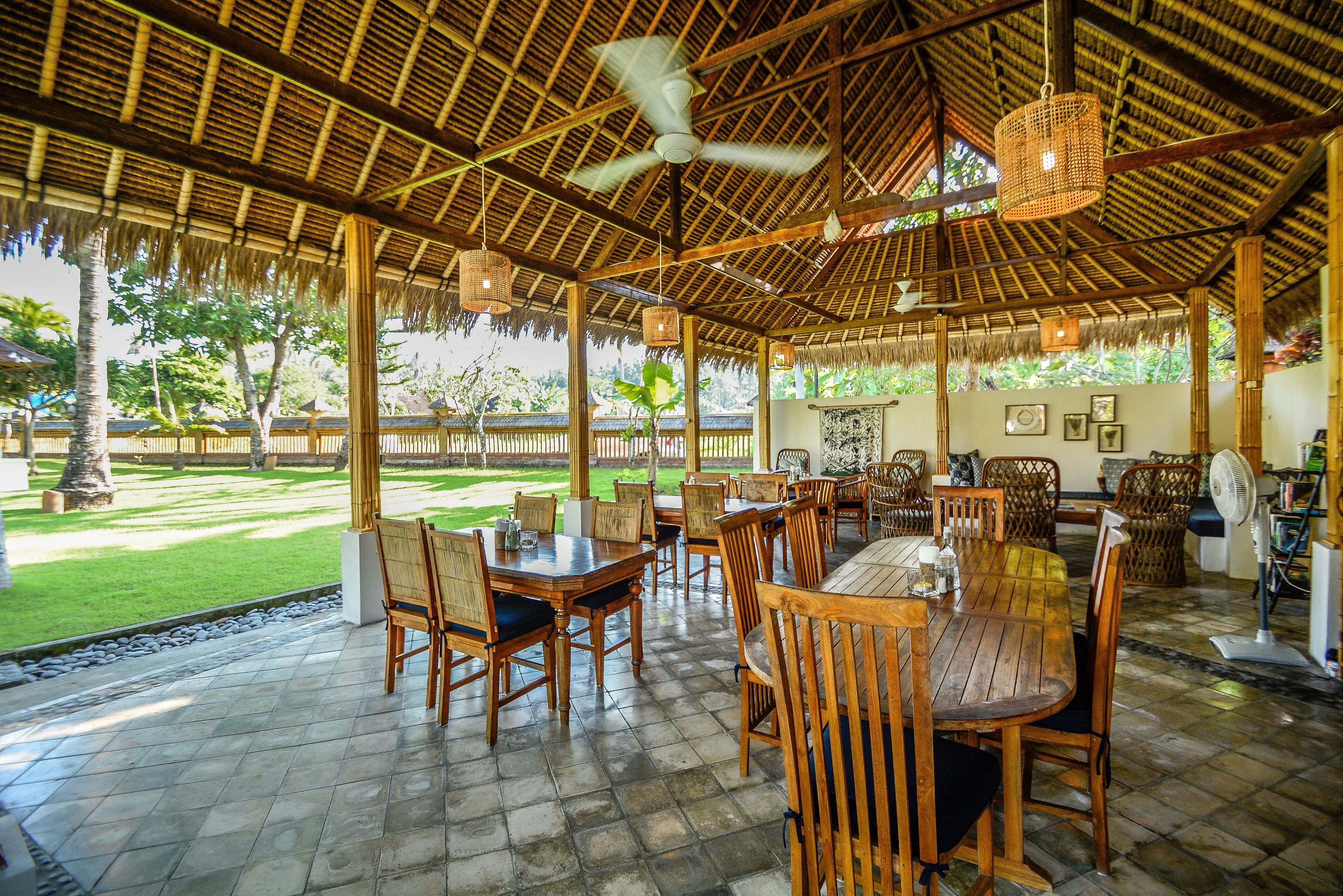 An outdoor dining area with wooden tables and chairs, a thatched roof, and a view of a well-maintained lawn.