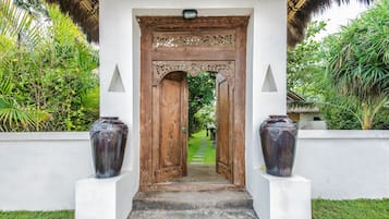 A wooden door with intricate carvings, flanked by two large vases on pedestals, leading to a pathway surrounded by lush greenery.