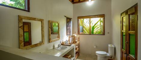 A bathroom with a white sink, two mirrors, a toilet, and a window with a view of greenery.