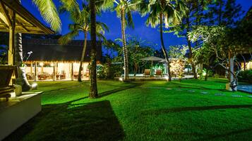Un espacio exterior bien iluminado con zona de comedor, palmeras y un edificio con techo de paja.