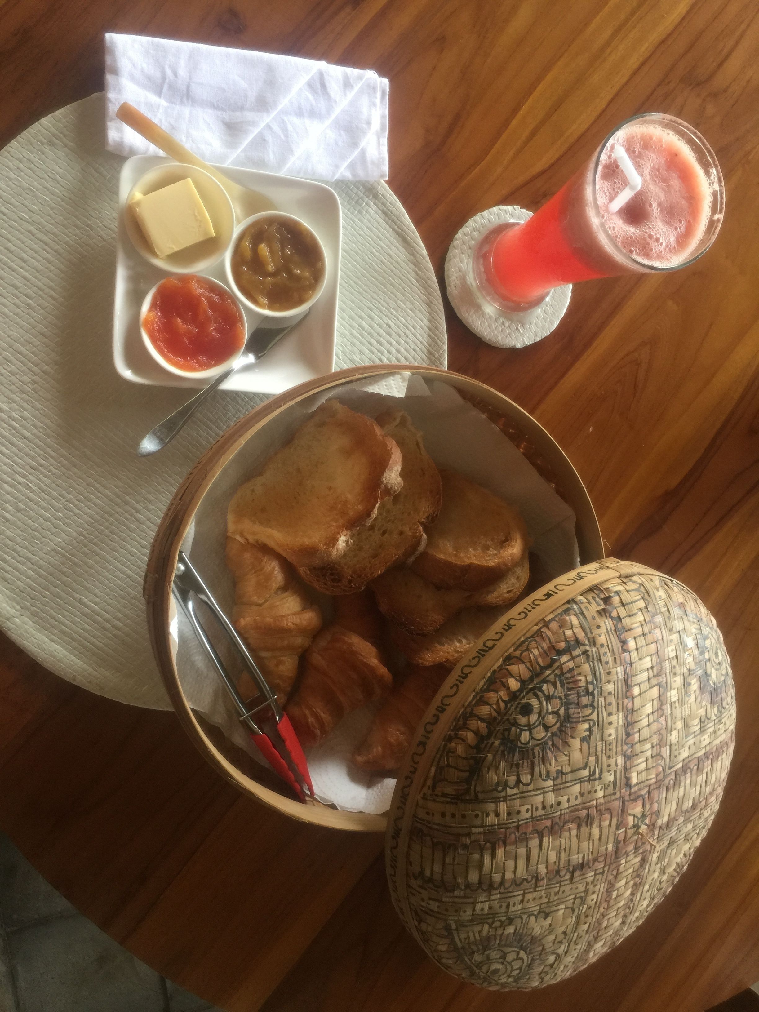 A table with a basket of bread rolls, a woven basket, a glass of red drink, and a plate with butter, chutney, and a sauce.