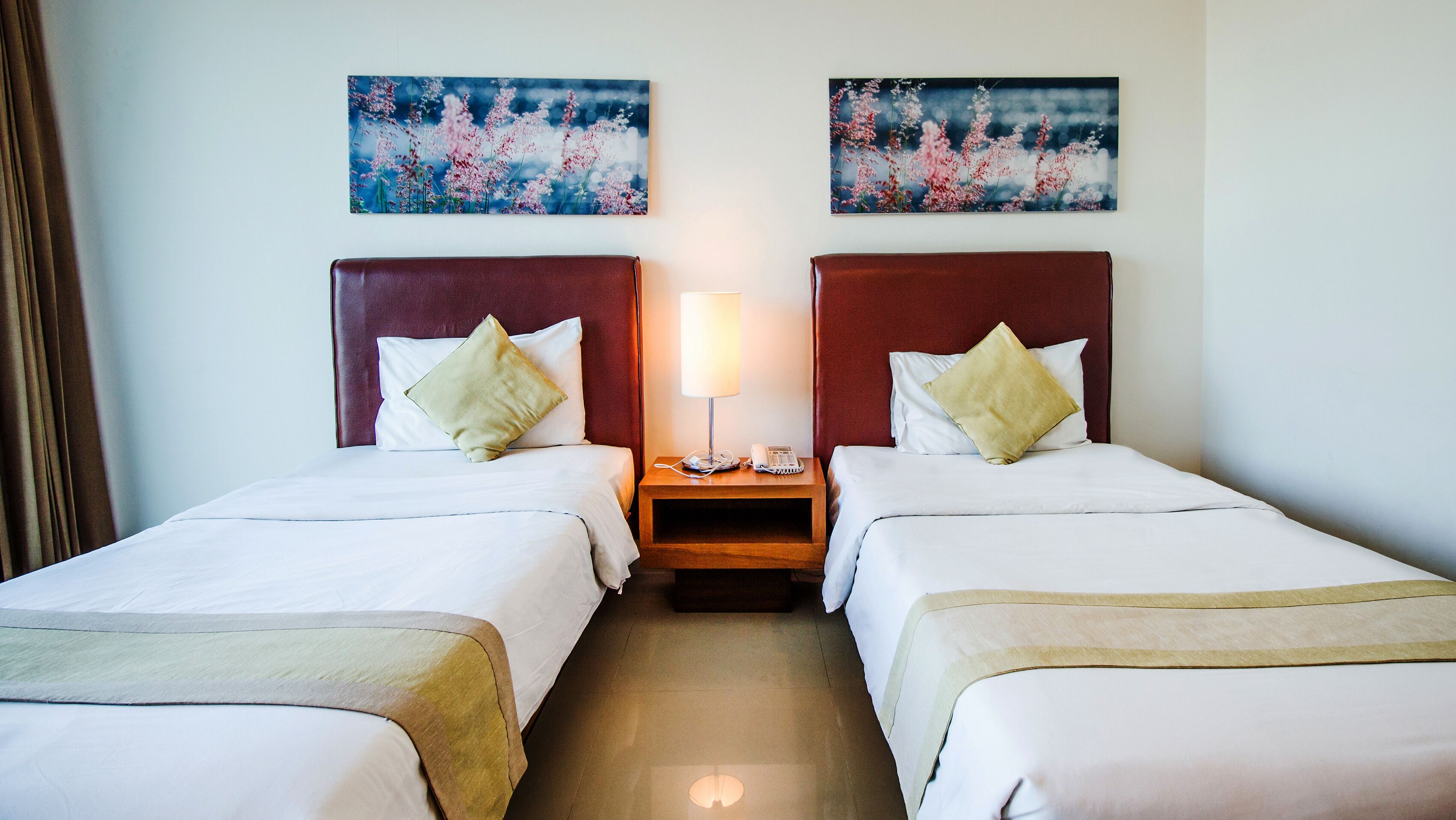 A hotel room with two beds, a nightstand with a lamp and phone, and two paintings on the wall.