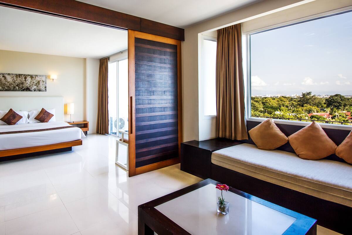A modern hotel room with a large bed, a sofa by the window, and a view of the city.