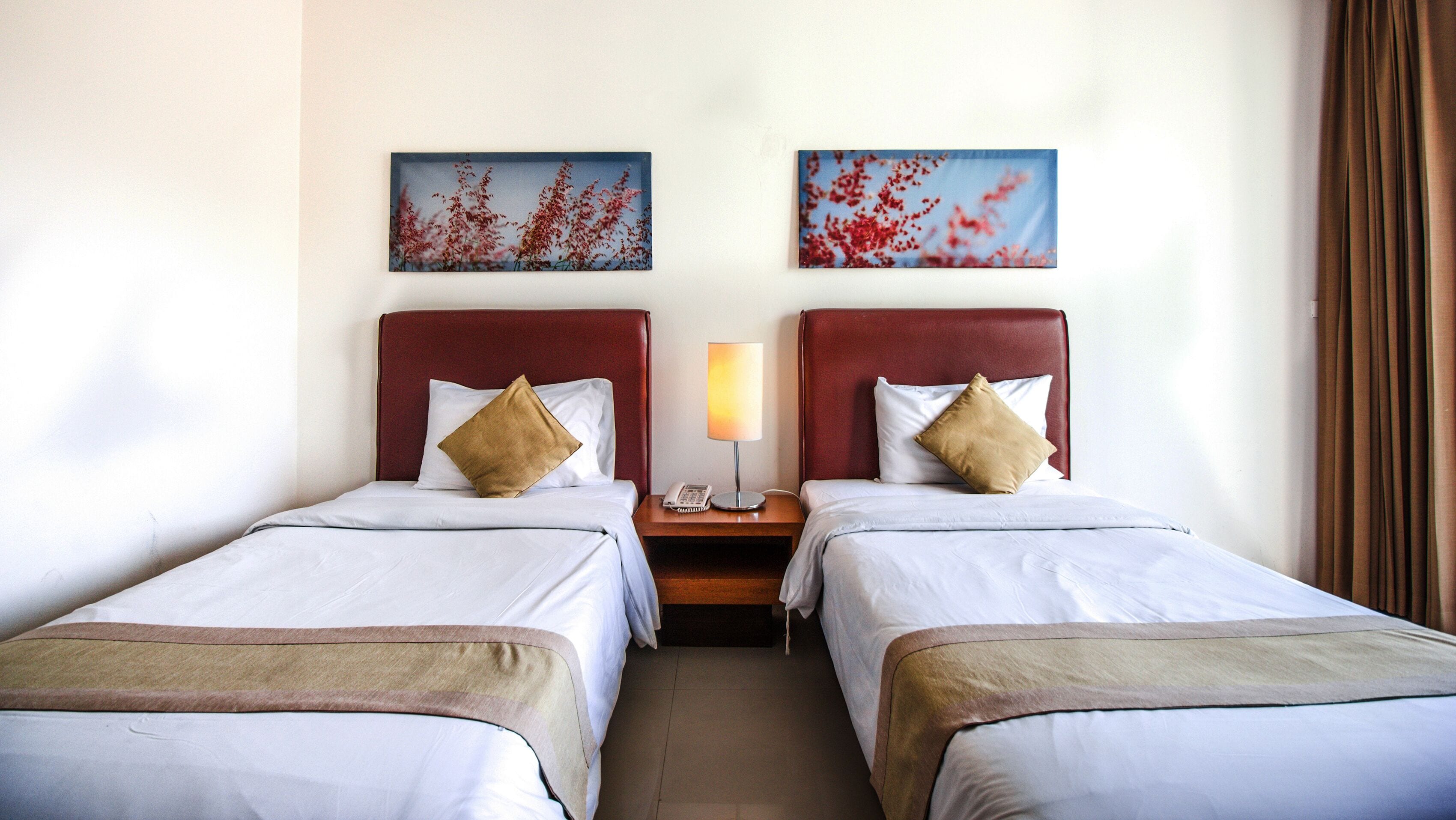 A hotel room with two beds, each with a red headboard, white bedding, and a beige throw. There are two paintings on the wall, a nightstand with a lamp, and a telephone between the beds.